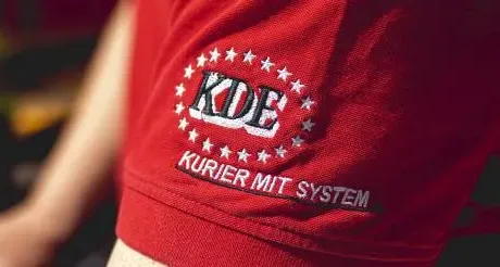 Der Fotograf hat hier den Ärmel der Dienstbekleidung eines Kuriers der KDE-Transport GmbH aufgenommen. Auf dem roten Shirt ist das KDE-Logo aufgestickt.
