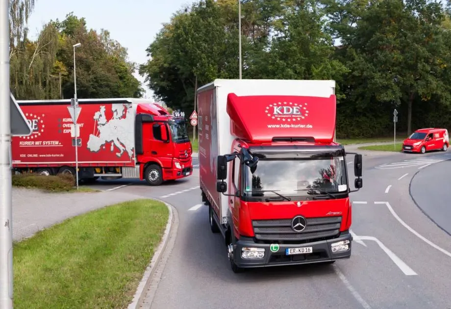 Die Aufnahme zeigt drei verschiedene Fahrzeuge der KDE Transport GmbH im Begegnungsverkehr an einer Kreuzung.