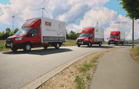 3 kleinere LKW der KDE Transport GmbH fahren auf einer Landstraße hintereinander. Im Hintergrund sieht man drei Windränder.