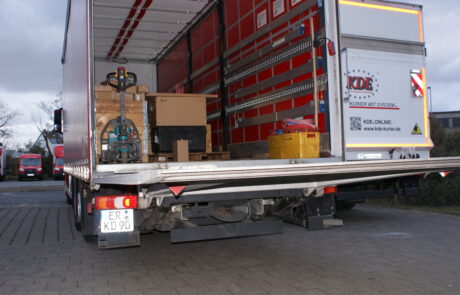 Das Foto zeigt zwei KDE-LKW von hinten. Eines der Hebebuehnenfahrzeuge ist geoeffnet und man sieht die geladene und gesicherte Ware.