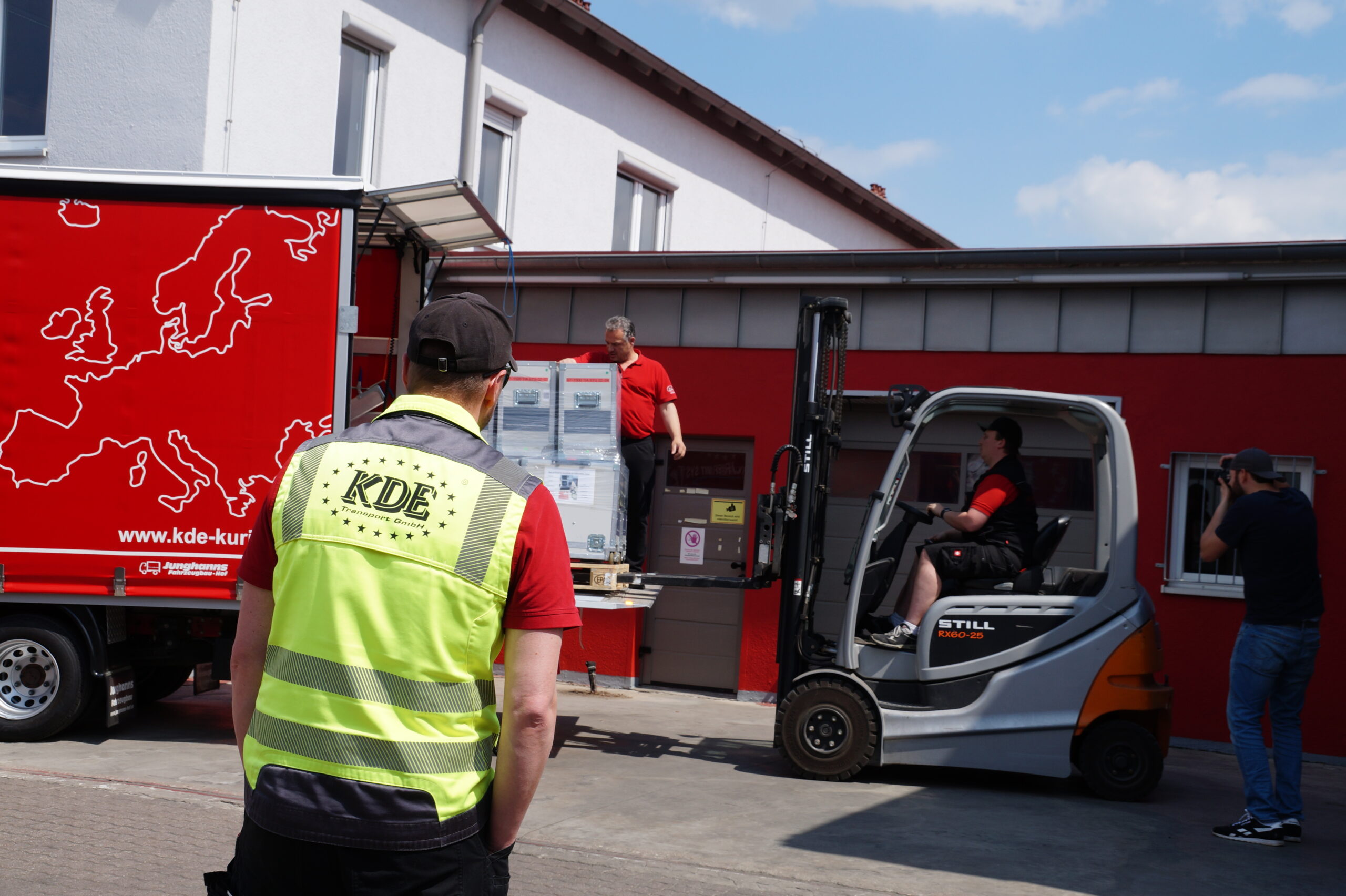 Das Foto zeigt einen Beladevorgang auf dem Gelände der KDE Transport GmbH. Ein Gabelstaplerfahrer stellt gerade eine schwere Palette auf der Hebebühne eines LKW ab. Ein Kurier überwacht den Vorgang auf der Hebebühne. Ein dritter Kurier in KDE-Sicherheitsweste sieht zu, ein Fotograf lichtet die Szene ab.