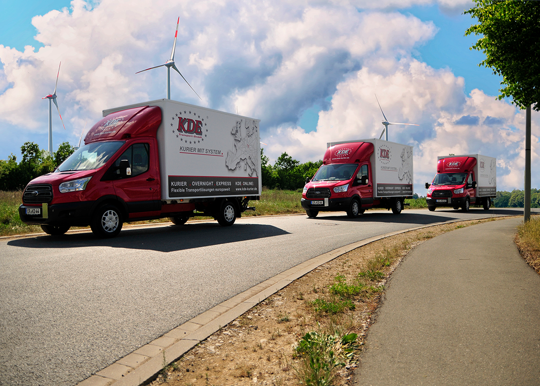Die Aufnahme zeigt drei LKW der KDE Transport GmbH auf Kurierfahrt. Im Hintergrund sieht man Windkraftraeder.